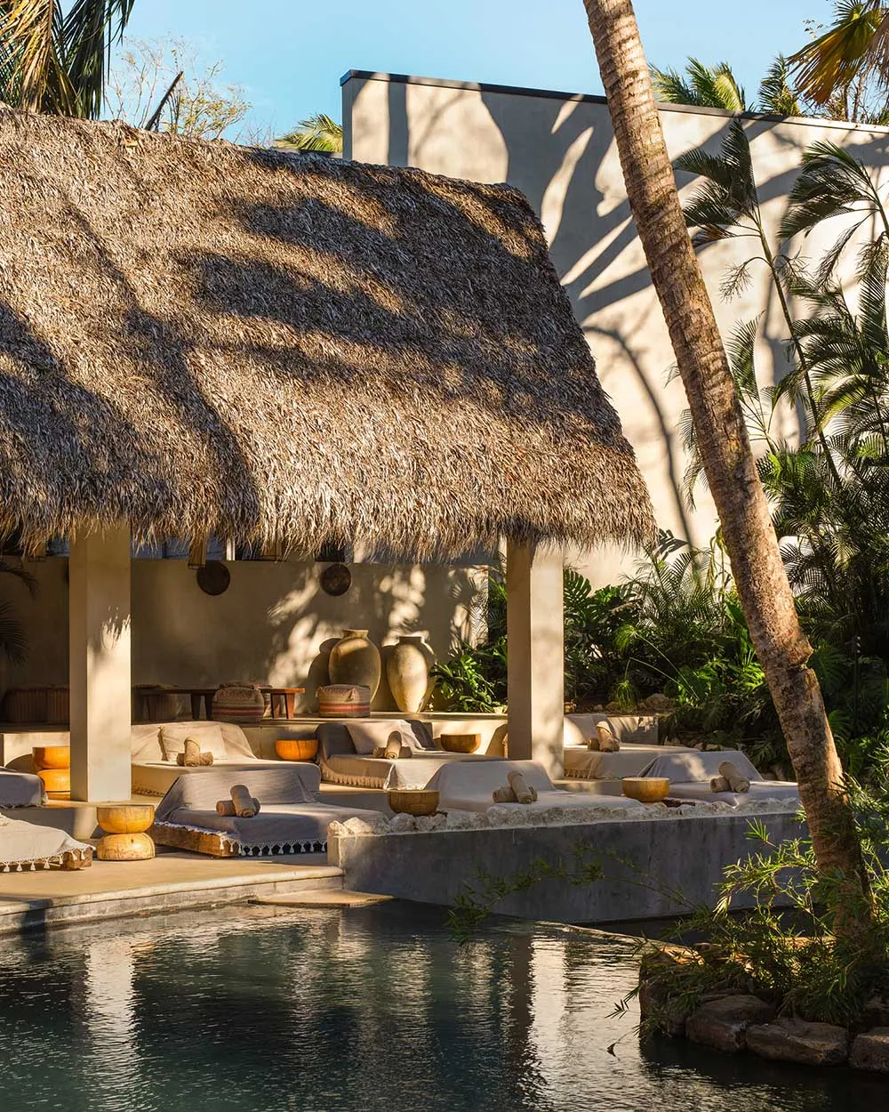 Luxurious outdoor daybed lounge with multiple white cushioned platforms under a large thatched roof, surrounded by palm trees and a reflecting pool.