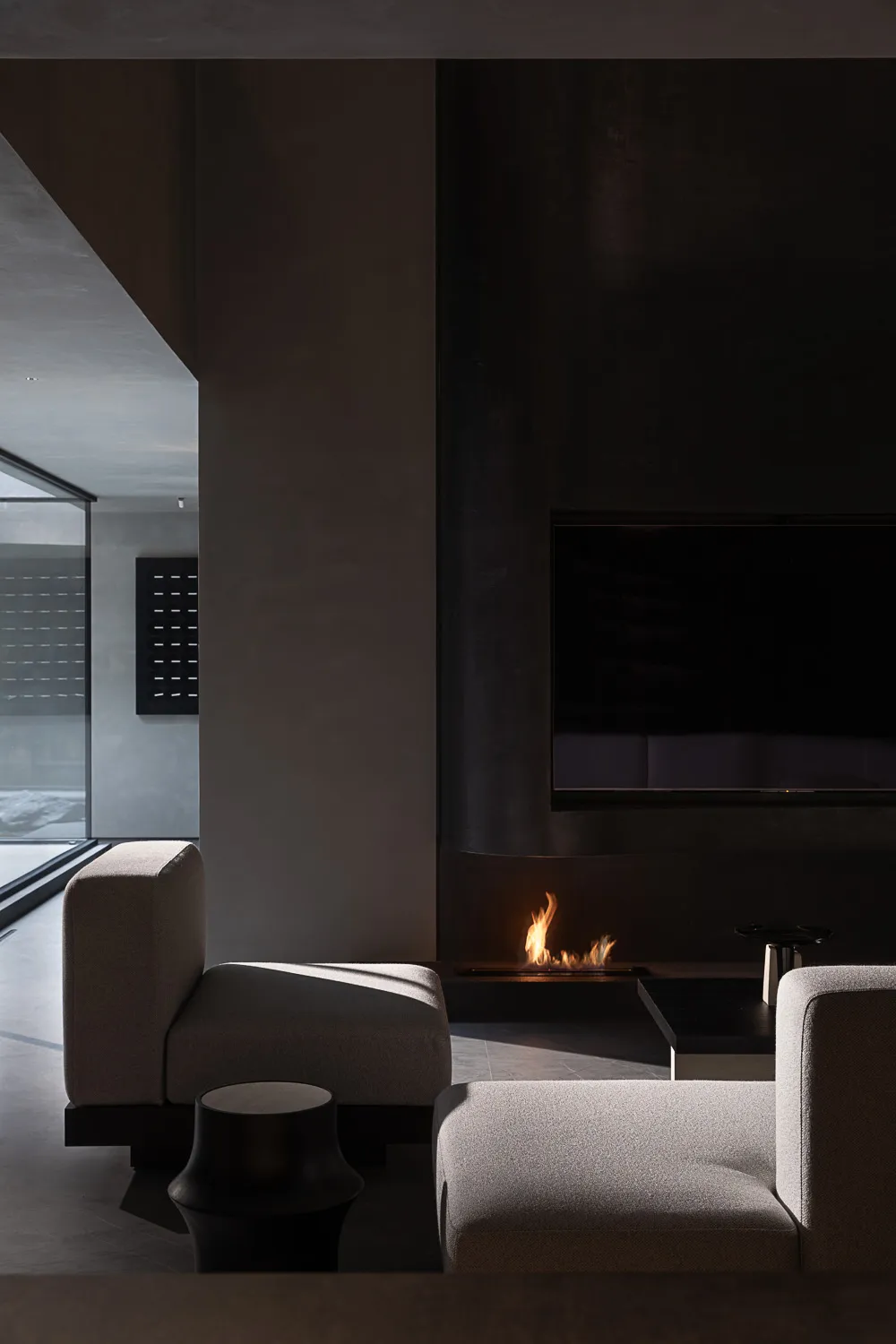 Modern minimalist living room featuring a dark concrete fireplace with burning fire, light grey modular sofas, and a black side table under dramatic natural lighting.