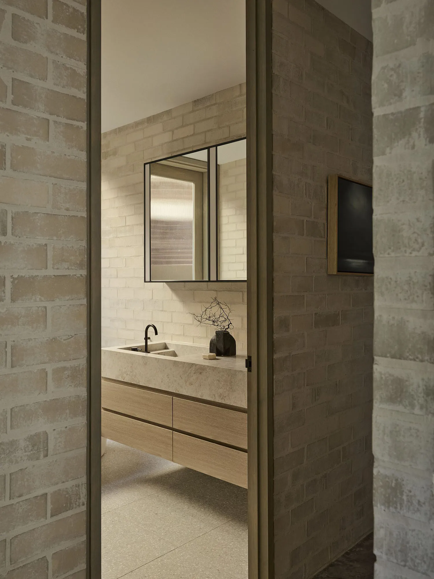 View into a modern brutalist bathroom with light travertine floating vanity, oak drawers, black matte faucet, and textured brick walls.