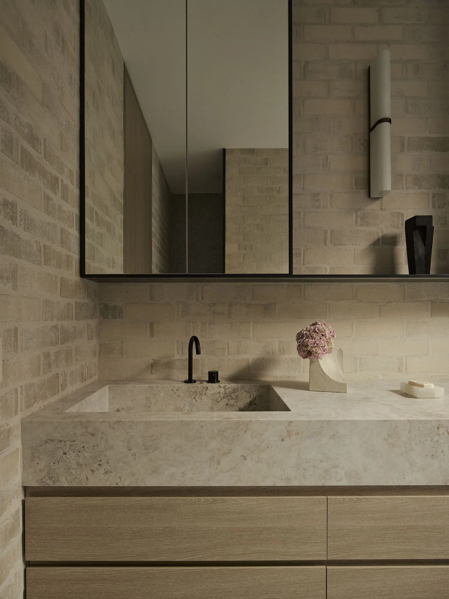 Modern bathroom featuring a solid travertine floating vanity with integrated sink, matte black faucet, light wood cabinetry, and textured beige brick walls with a large frameless mirror.