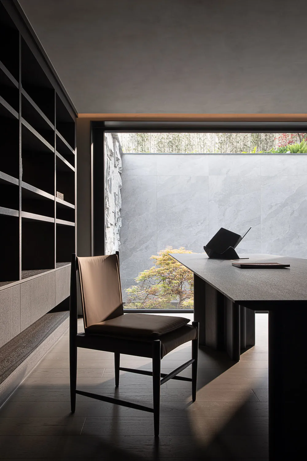 Modern minimalist home office featuring a dark walnut desk, brown leather chair, built-in shelving, and a large window overlooking a tranquil garden with stone wall.