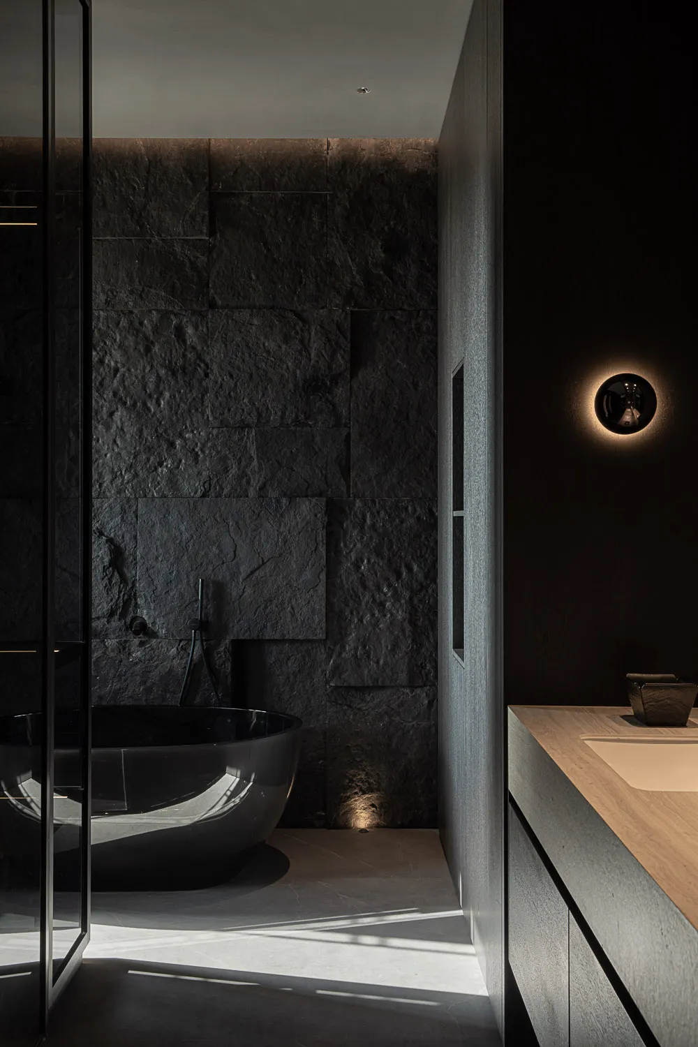Modern dark minimalist bathroom featuring a glossy black freestanding tub against a textured natural stone wall, paired with a light wood floating vanity and dramatic wall sconce lighting.