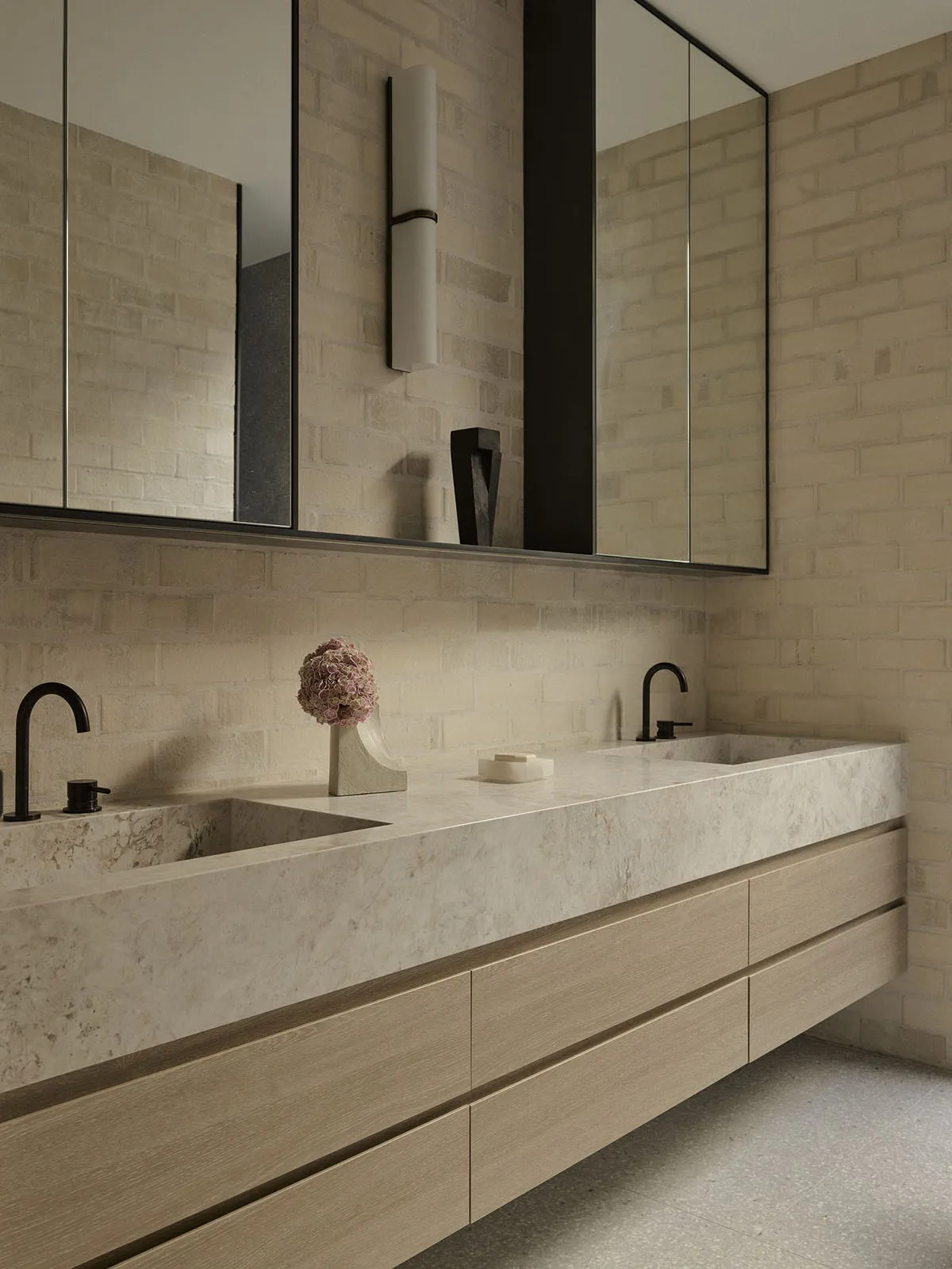 Modern minimalist bathroom with a long floating double vanity made of light marble, matching marble sinks, matte black faucets, large mirrors with black frames, and a beige brick accent wall with vertical wall sconce.