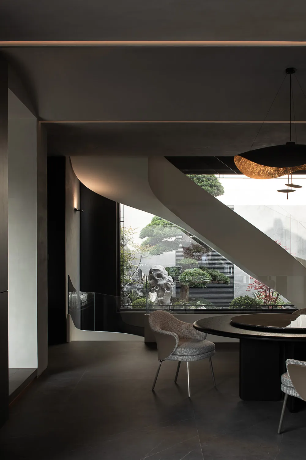 Modern minimalist dining area featuring a dramatic curved concrete wall, dark micro-cement floors, round dark dining table, textured bouclé chairs, and a large window overlooking a Japanese-inspired bonsai and rock garden.
