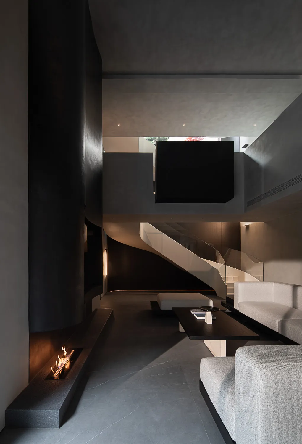 Modern double-height living room featuring a dramatic black micro-cement fireplace, floating staircase, light grey modular sofa, and dark coffee table under high ceilings.