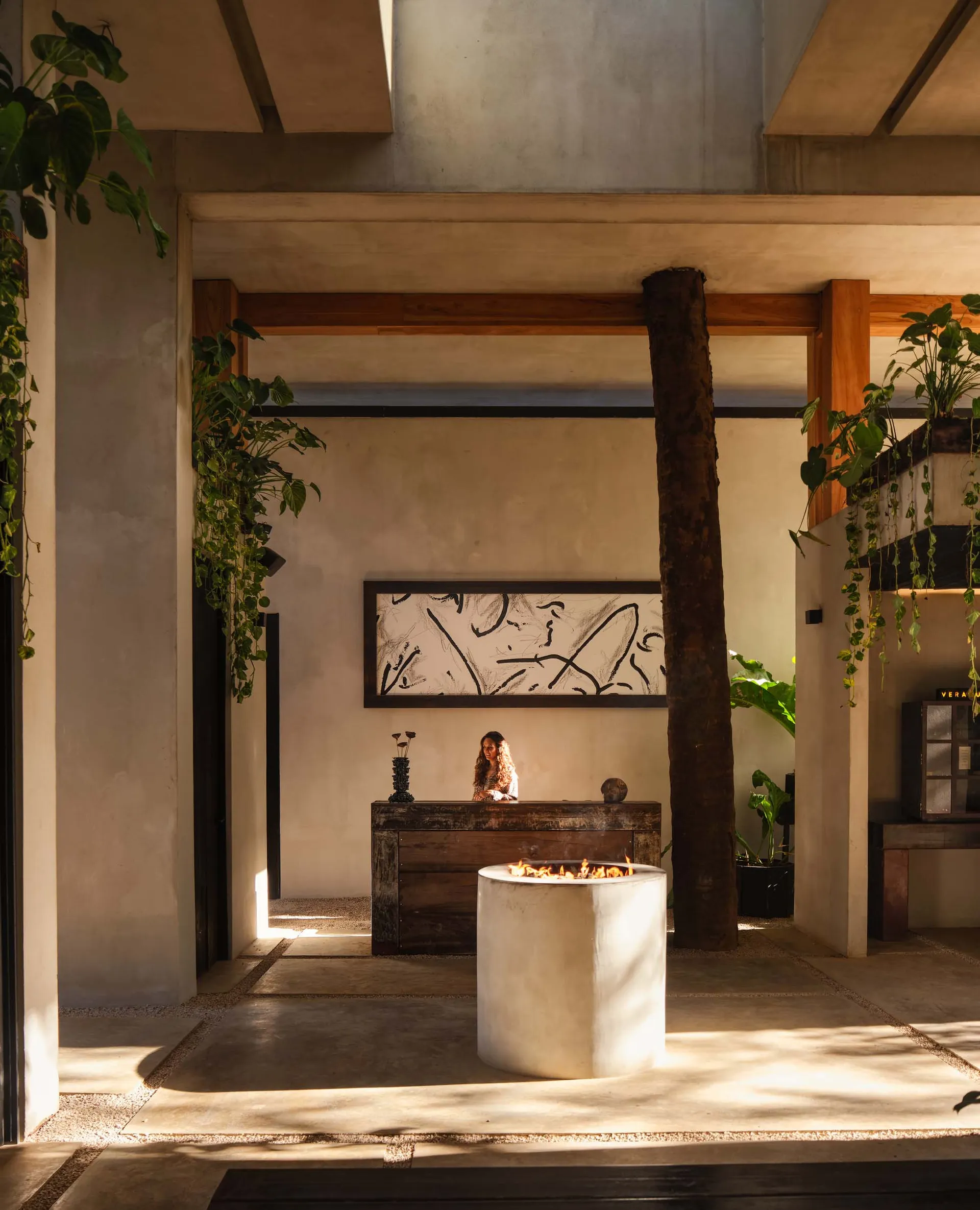 Modern tropical lobby interior with concrete fire pit in foreground, dark wood reception desk, large abstract artwork, wooden columns, and abundant hanging plants under bright natural light.