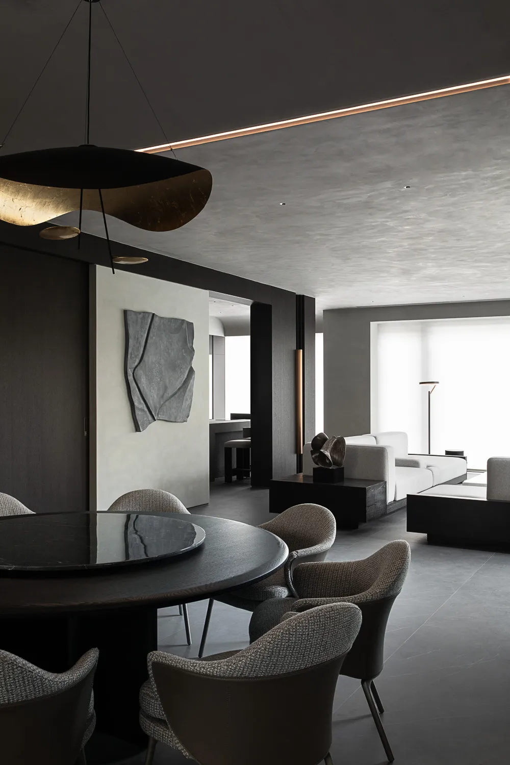 Modern minimalist dining area featuring a large round dark wood table, six textured boucle and leather chairs, an oversized black and gold sculptural pendant light, concrete walls, and a view into a light-colored living room with white sofas.