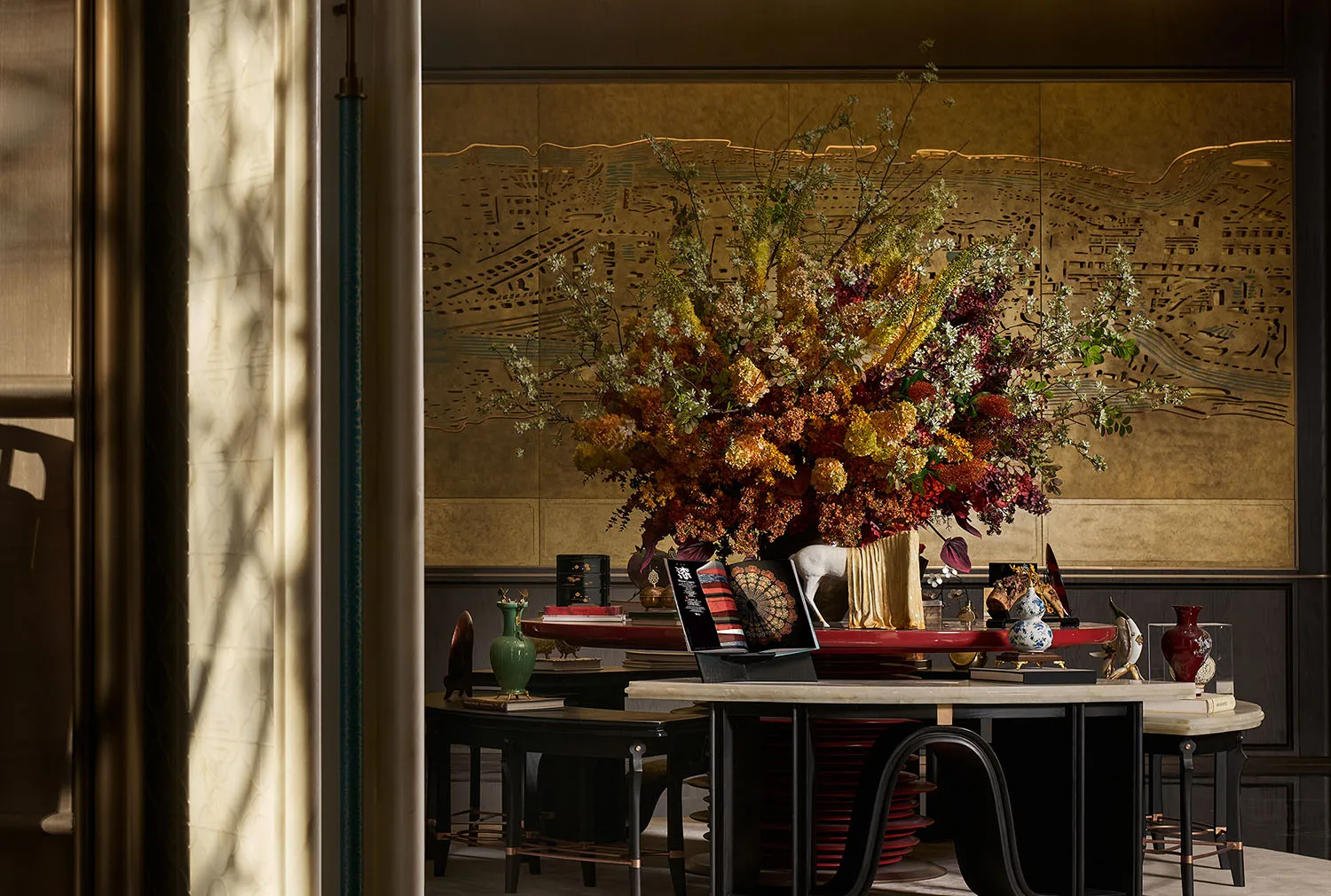 Luxurious entryway console table featuring a massive autumnal floral arrangement with dried flowers and branches, set against a hand-painted gilded Chinoiserie screen, with black lacquered furniture and porcelain accents.
