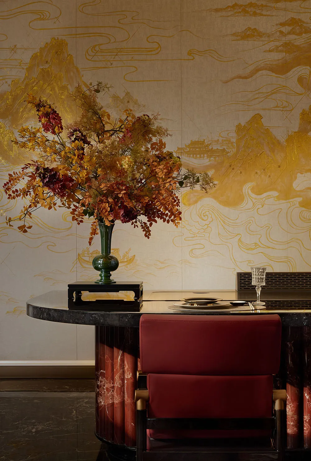 Round black marble dining table with red velvet upholstered chair, large autumnal floral arrangement in a green vase, and dramatic golden Chinese mountain-and-cloud mural on the wall.