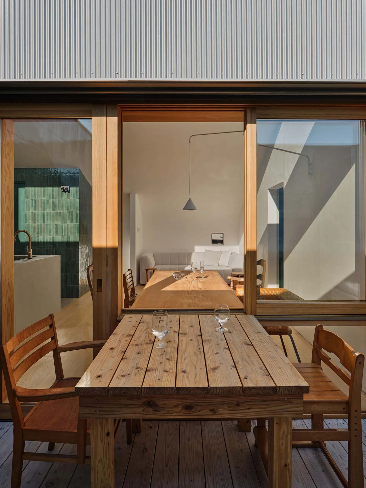 Wooden dining table and chairs on a timber deck with large open sliding doors revealing a minimalist living room and kitchen in the background