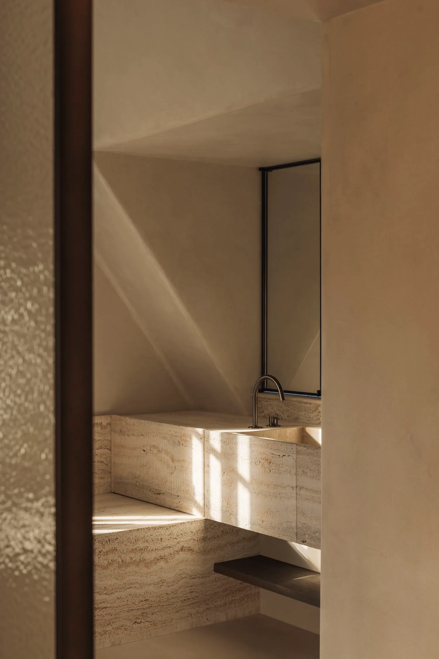 Modern minimalist bathroom featuring a floating travertine vanity with integrated sink, dramatic natural light casting strong shadows on textured stone walls and a frameless mirror.