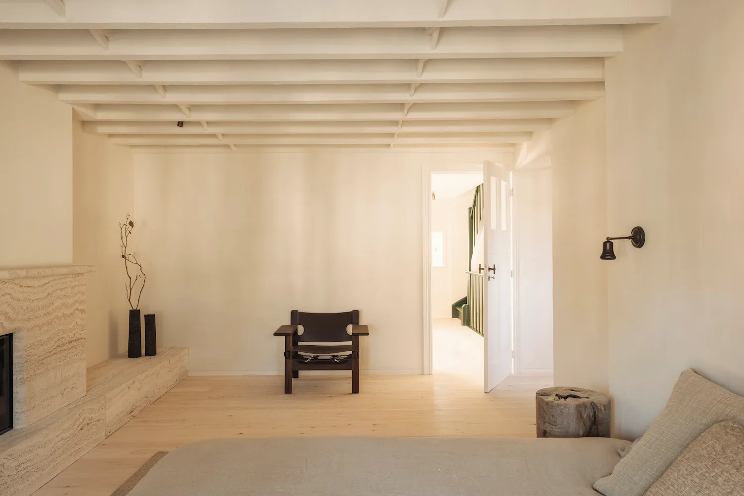 Minimalist living room featuring a travertine fireplace, dark oak sling chair, light wood floors, and exposed ceiling beams with an open doorway leading to another room.