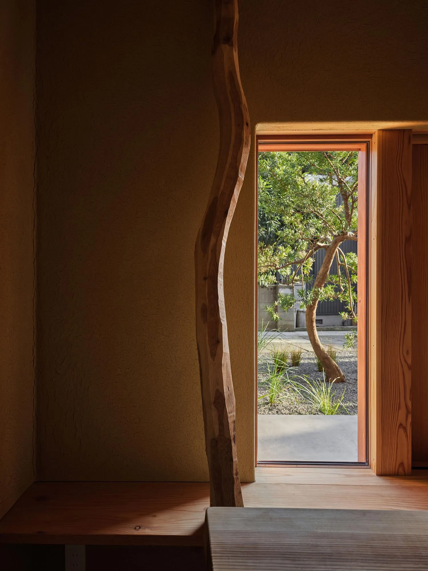 Interior view of a minimalist wooden space with a large organic-shaped wooden frame opening to a Japanese garden with a twisted pine tree and gravel landscaping.