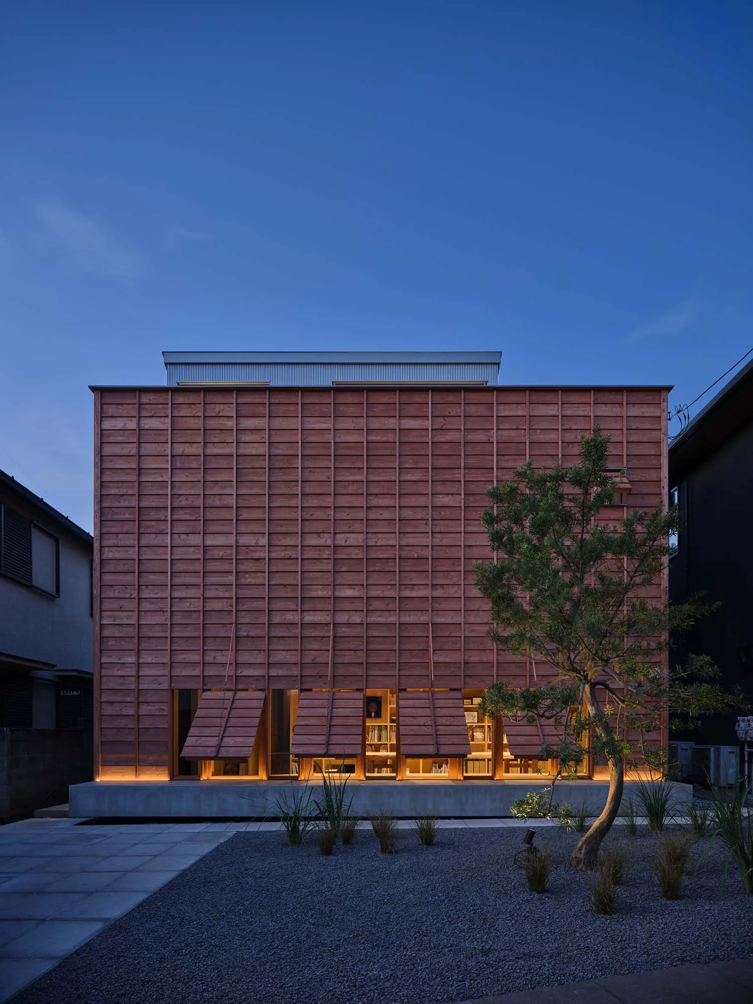 Modern two-story cedar wood house with horizontal wooden slats and warm-lit folding wooden awnings at dusk, surrounded by a minimalist gravel garden with a small pine tree.