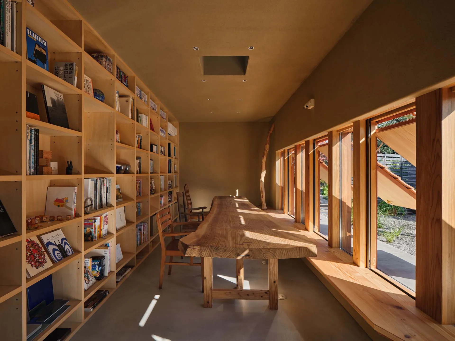 Long solid wood live-edge dining table in a bright wooden library room lined with tall built-in bookshelves and large windows letting in natural sunlight