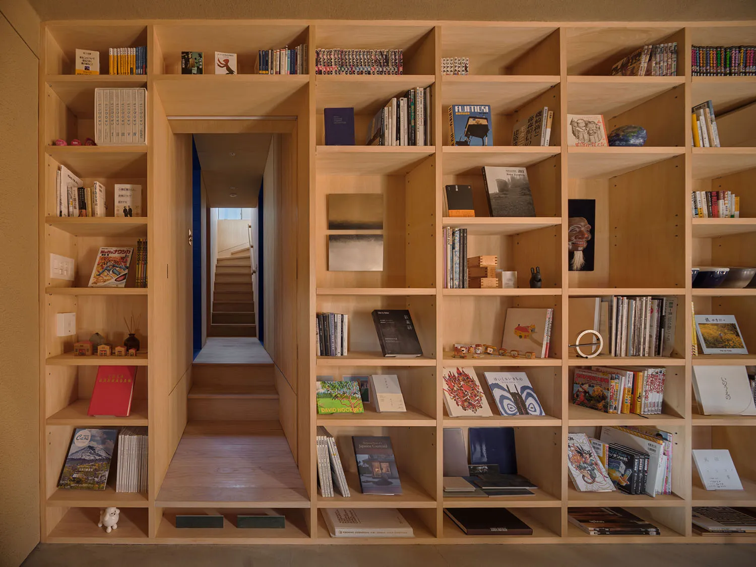Built-in light oak bookshelf wall featuring an integrated wooden staircase and doorway leading to another level, filled with books, magazines and decorative objects.