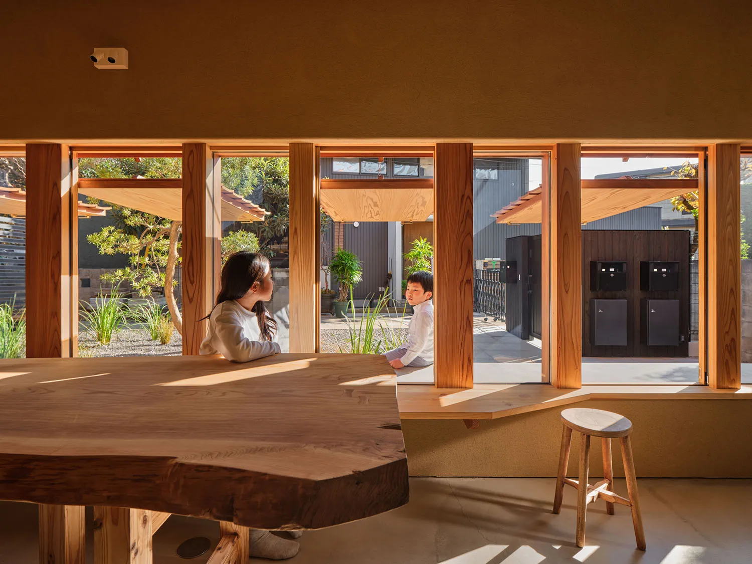 Two children sitting by a large solid wood live-edge dining table in a sunlit room with floor-to-ceiling wooden windows overlooking a Japanese-style courtyard garden.