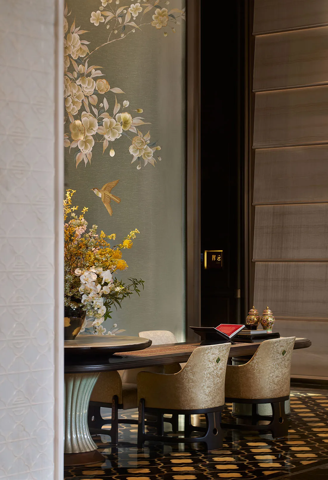 Luxurious Chinese-style dining room featuring a large hand-painted floral and bird mural in sage green and cream tones, a round dark wood dining table with a fluted jade-like base, and upholstered velvet dining chairs on a patterned dark floor.