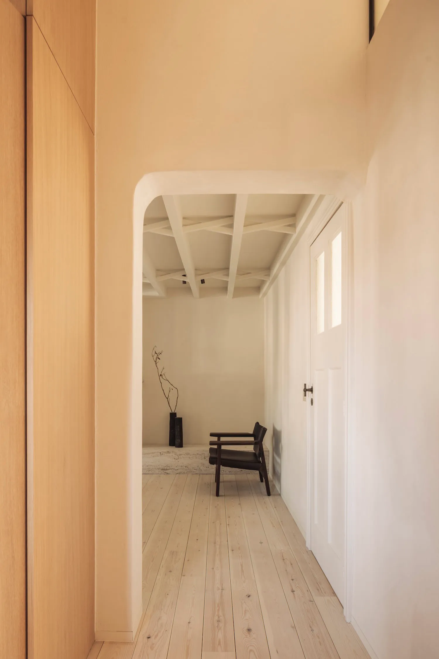 View down a minimalist hallway with light wood flooring, white walls, a dark wood armchair, and distinctive white sculptural ceiling beams.