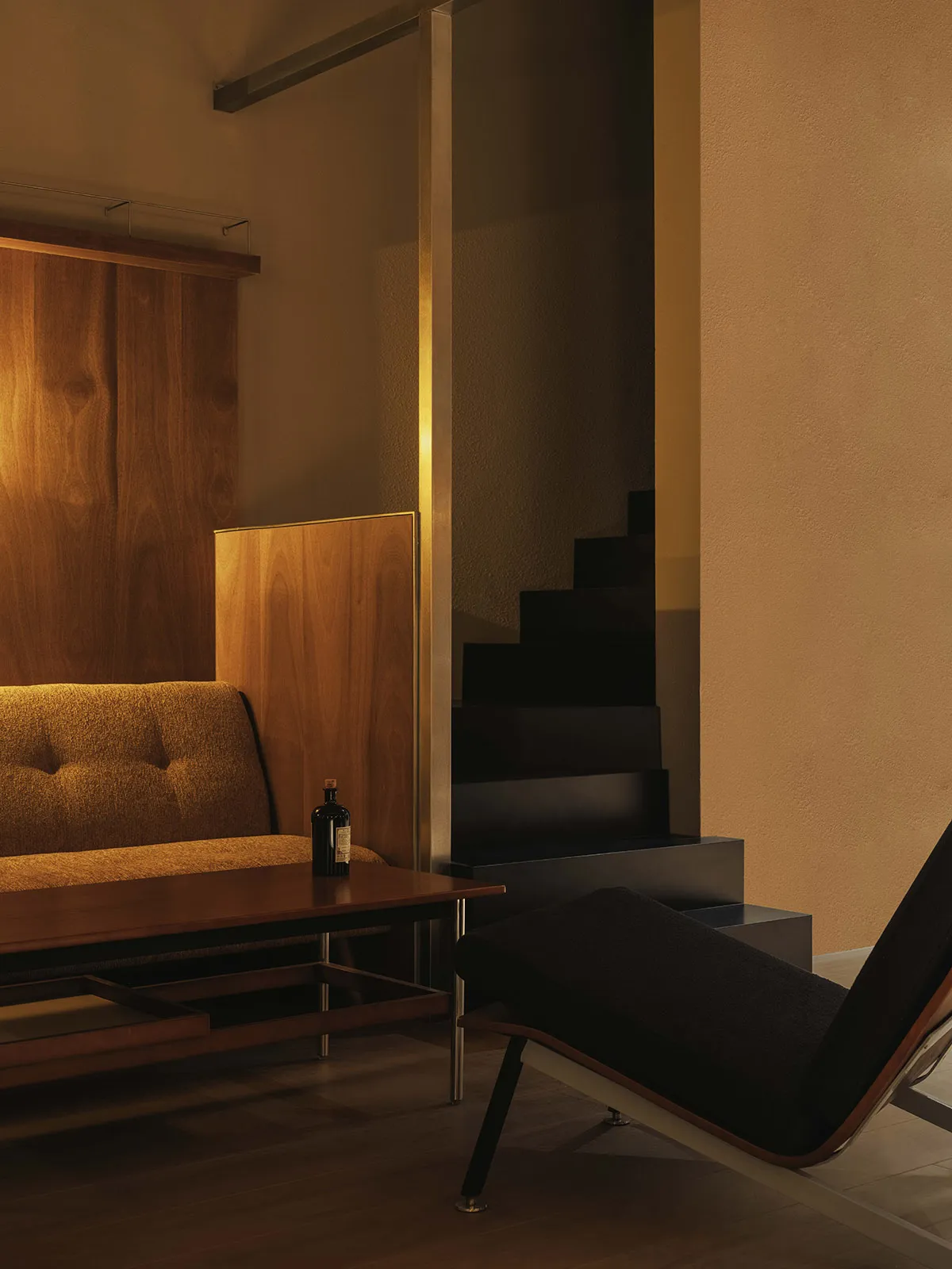 Modern living room corner featuring a mustard-yellow bouclé sofa, walnut wood paneling, a dark wooden coffee table, a black upholstered chaise lounge, and a black floating staircase in the background under warm lighting.