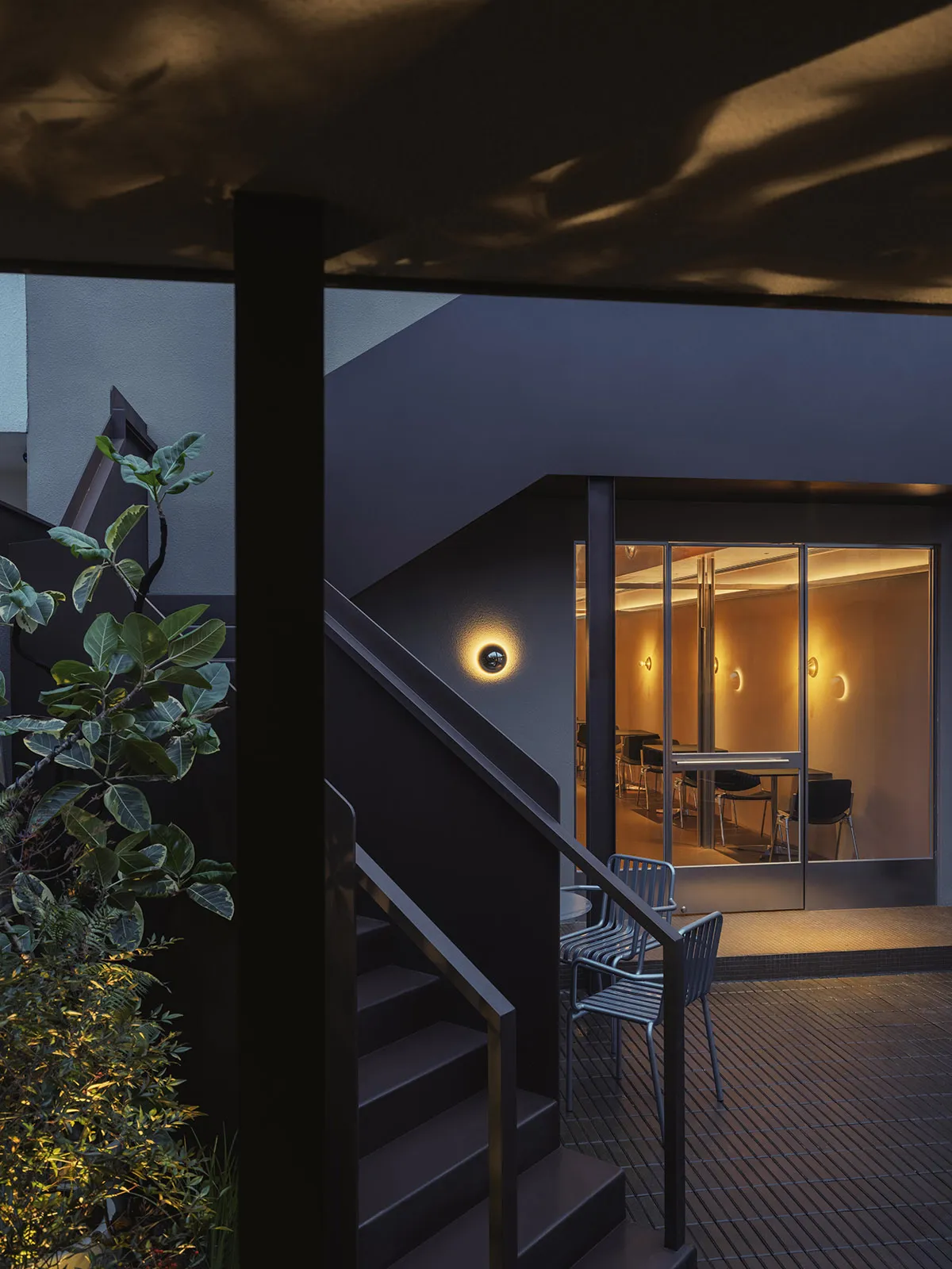 Night view of a modern dark-painted courtyard staircase with blue metal chairs on a wooden deck, leading to a warmly lit glass-walled dining room.