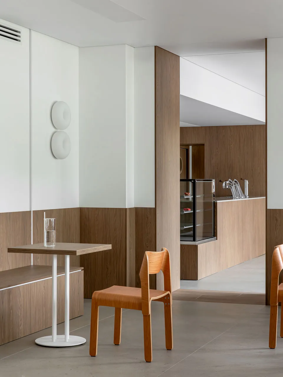 Modern minimalist cafe interior featuring light oak wood paneling on lower walls, white upper walls, a small square oak table with white base, and a bentwood oak chair.