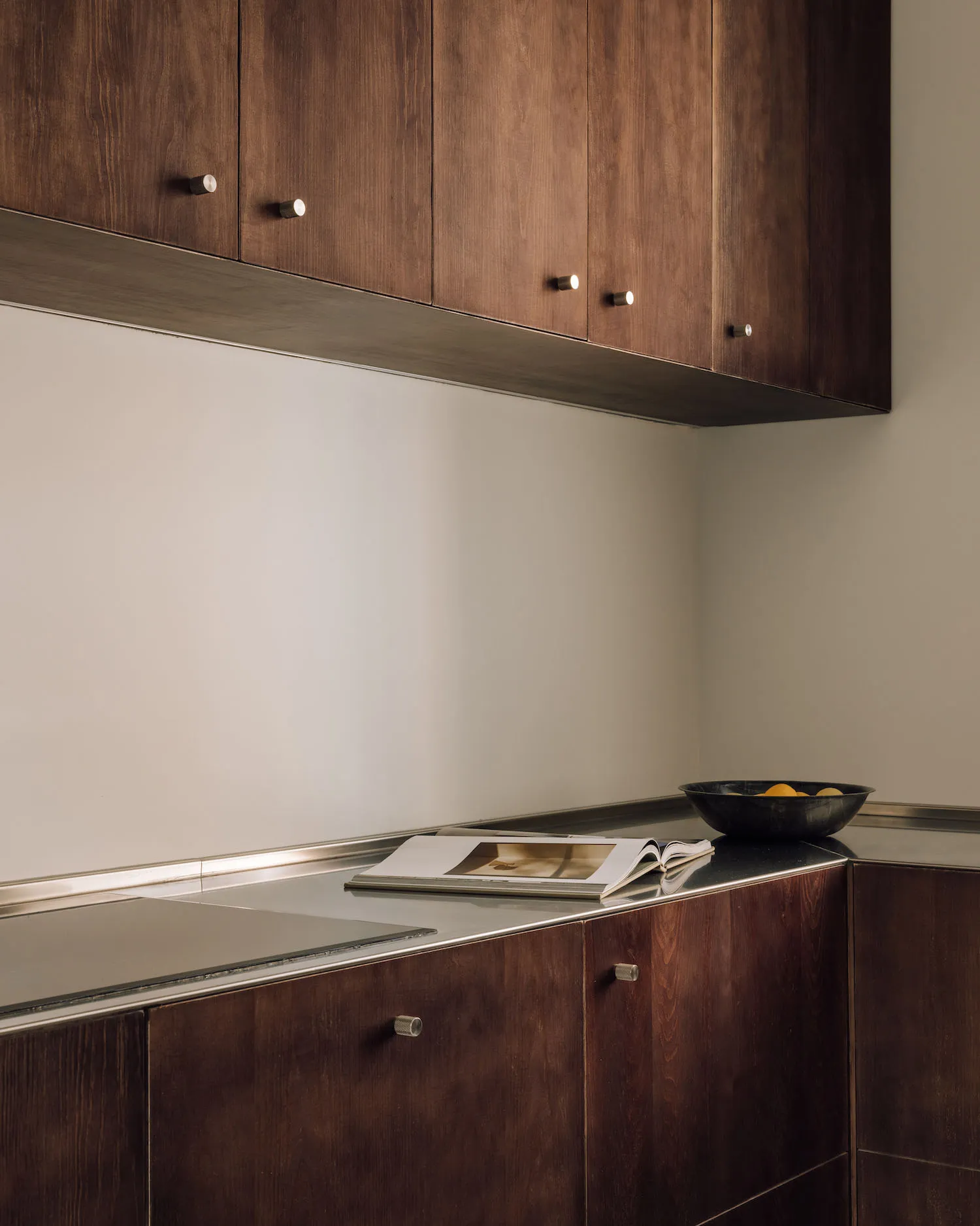 Modern minimalist kitchen corner featuring dark walnut upper and lower cabinets with metallic handles, stainless steel countertop, and a black bowl with lemons placed on the counter.