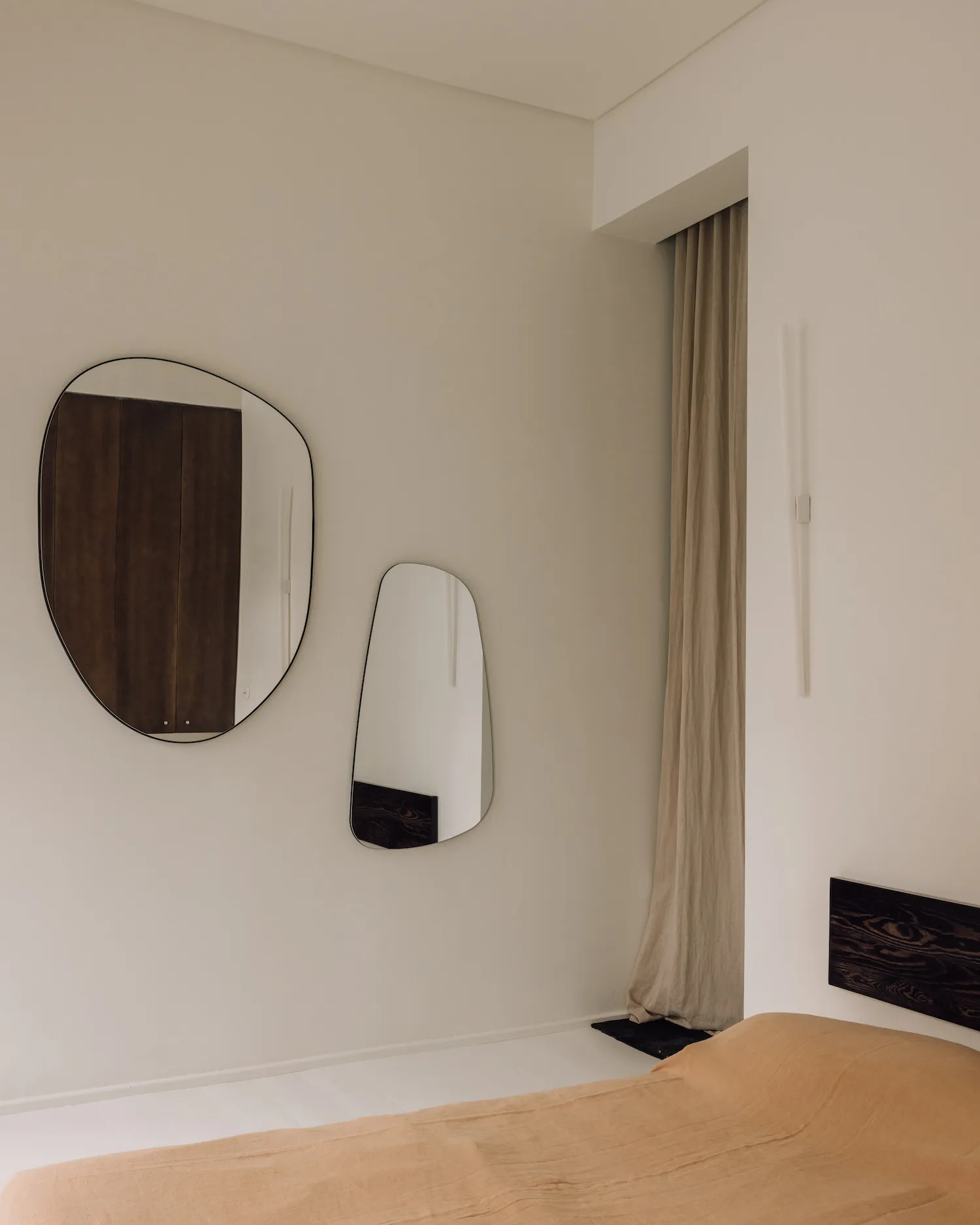 Minimalist bedroom corner featuring two asymmetrical mirrors with black walnut frames, beige linen curtains, and a walnut headboard against off-white walls.