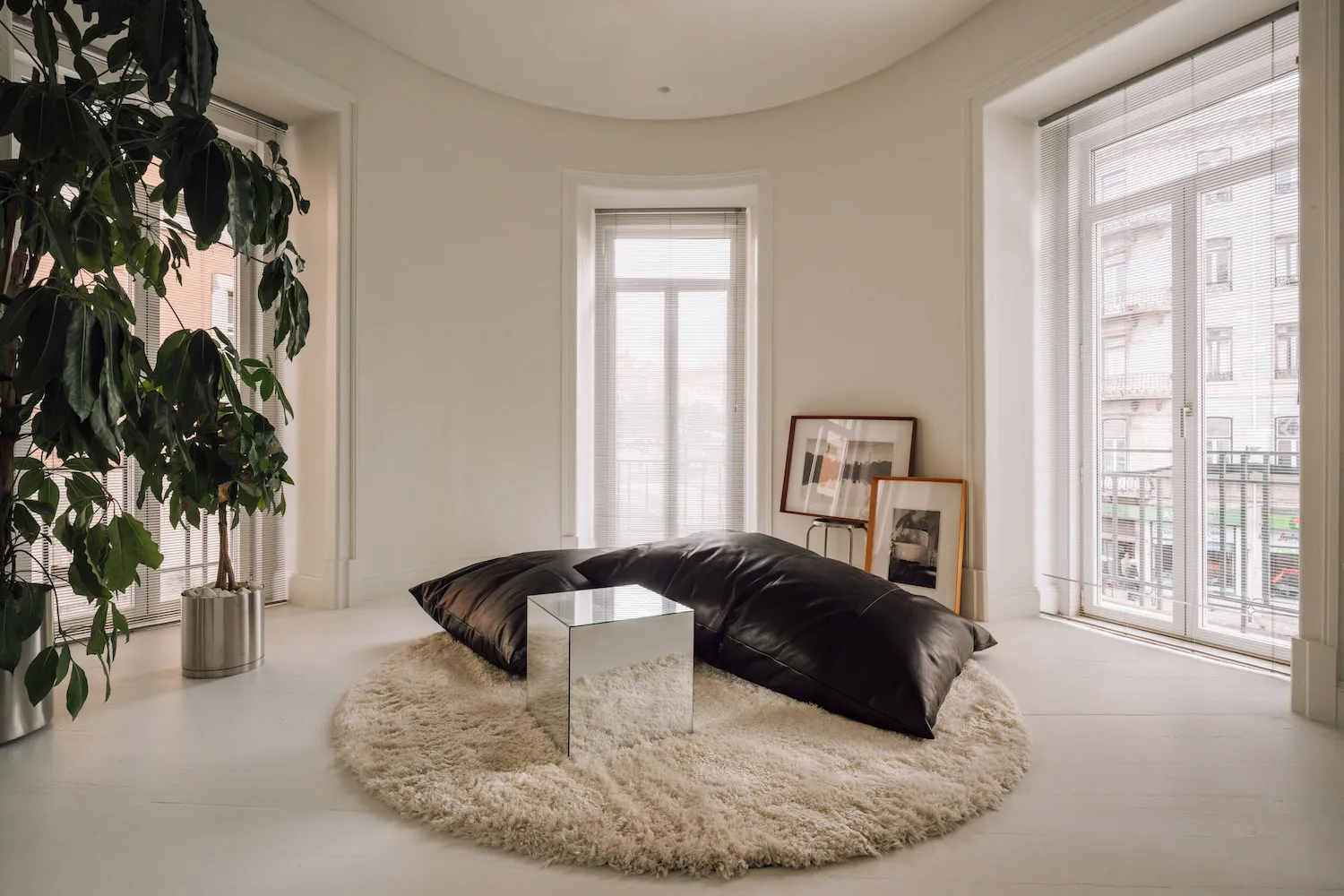 Bright minimalist living room with curved white walls, large windows with blinds, a circular cream sheepskin rug, black leather floor cushions, a mirrored acrylic side table, and a tall potted fiddle-leaf fig plant.