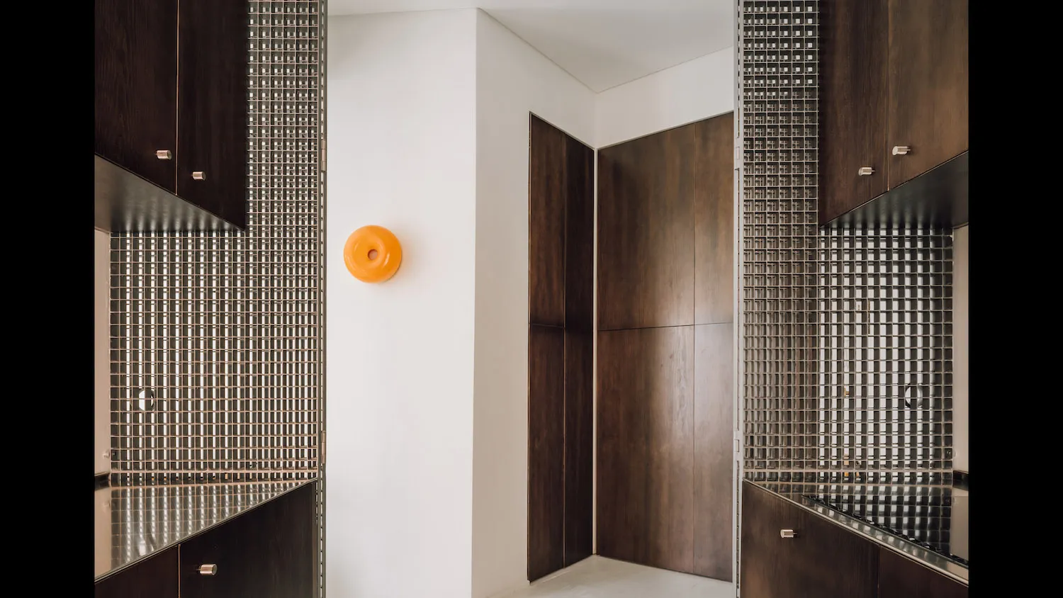 Modern minimalist entryway featuring dark wood cabinetry, silver mosaic tile walls, white walls, and a bright orange circular wall light fixture.