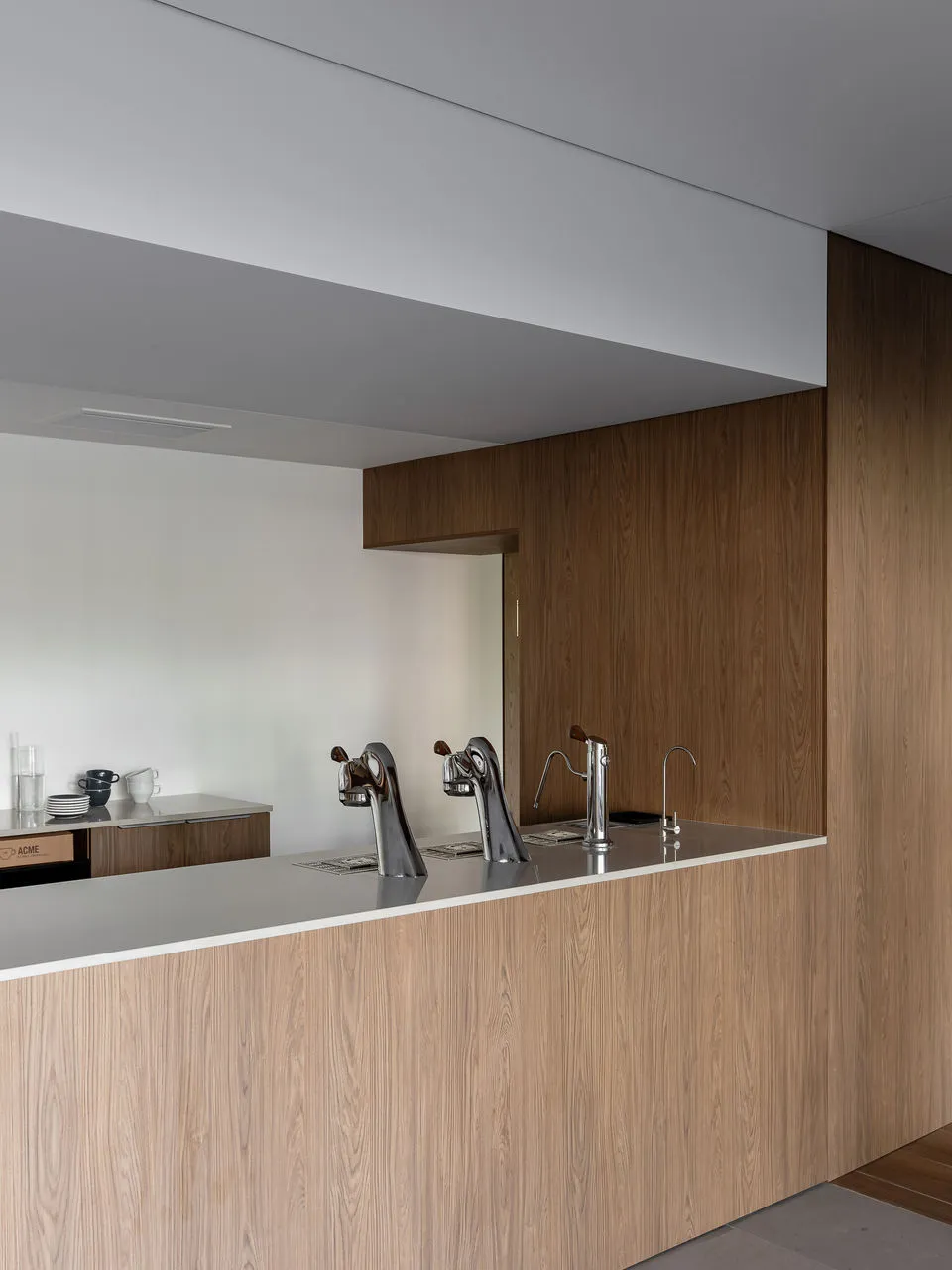 Modern minimalist kitchen island in light oak with white countertop and three sculptural chrome faucets against a white wall.