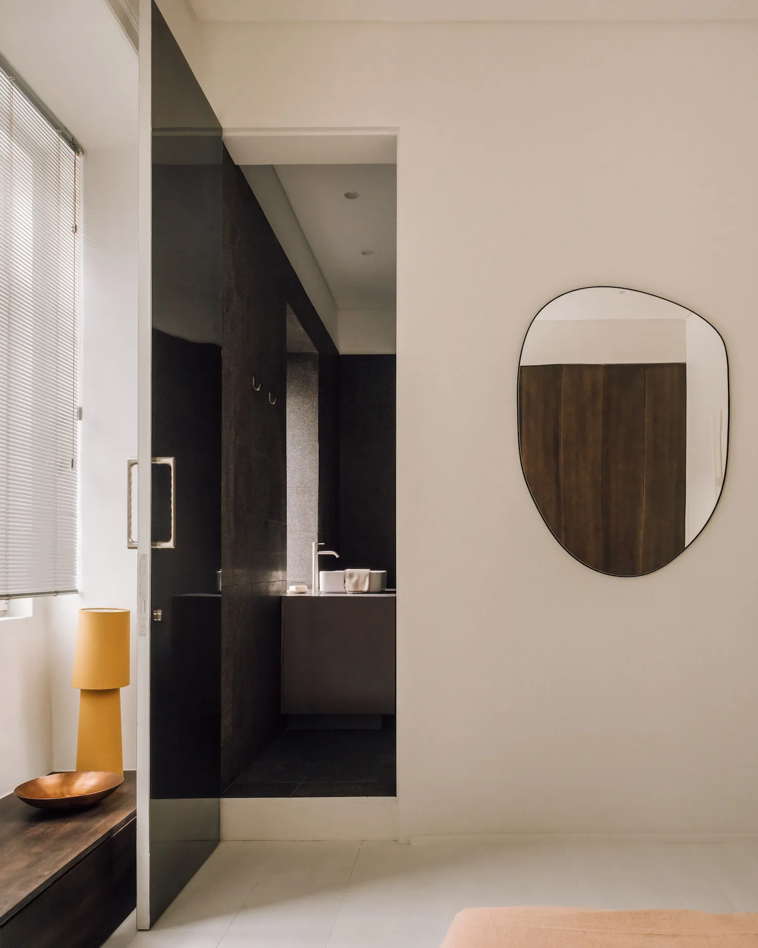 View from a bright room into a dark oak bathroom featuring an asymmetrical organic-shaped mirror, floating vanity, and warm yellow table lamp.