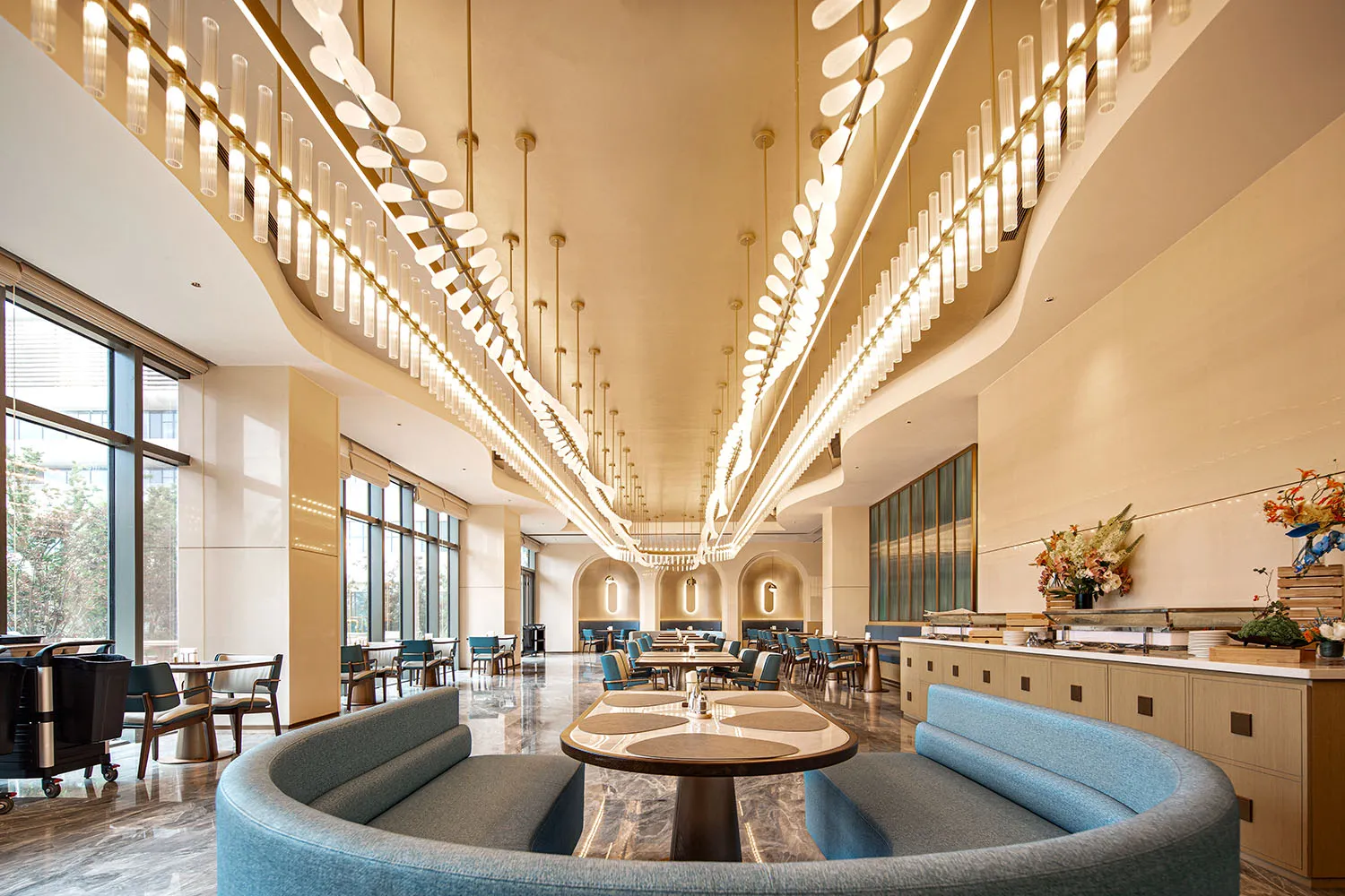 Modern luxury hotel breakfast room with curved blue fabric banquette, round marble dining table, dramatic long pendant lights, and polished marble floor.