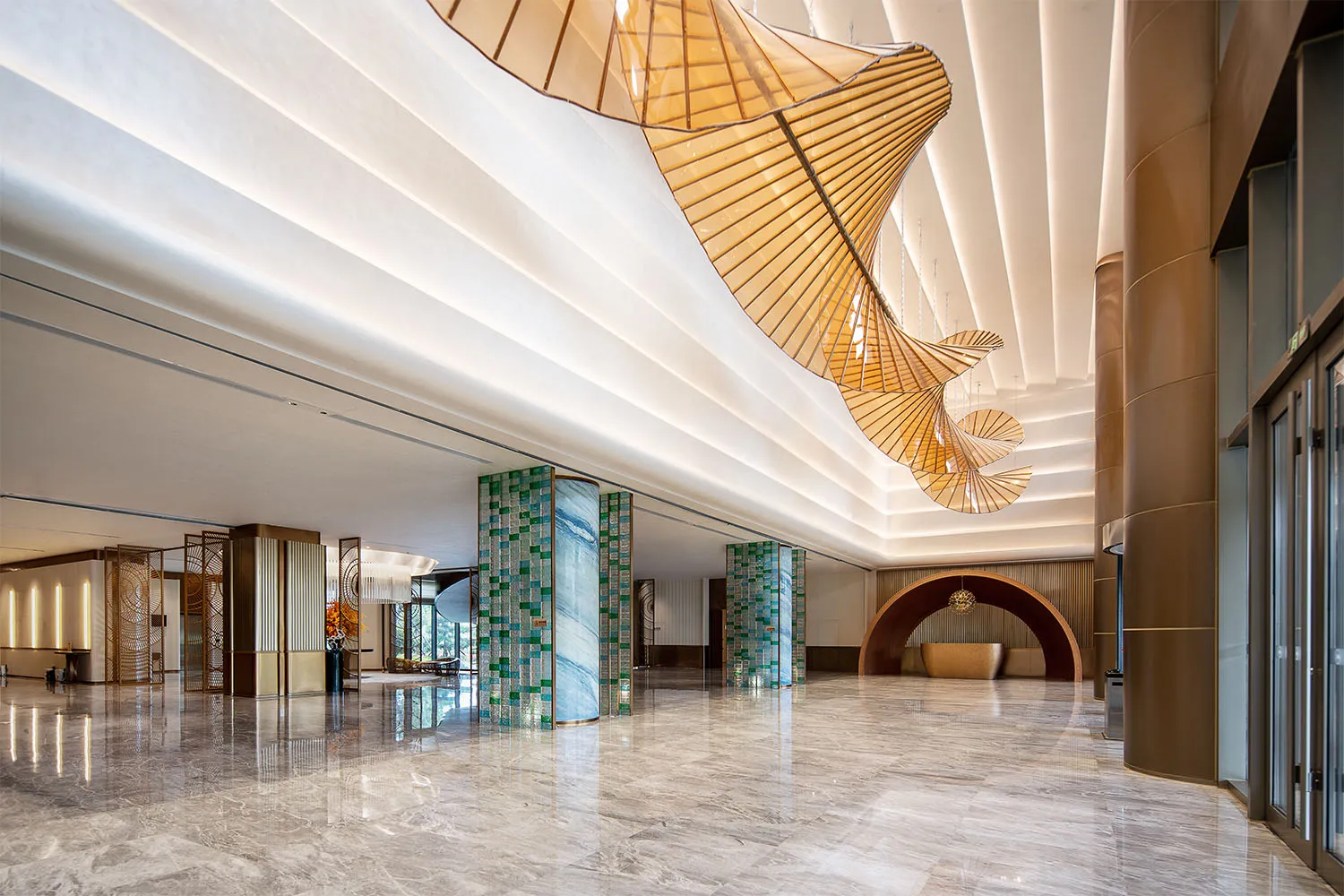 Modern luxury hotel lobby interior with polished marble floors, green marble pillars, and a massive undulating wooden chandelier hanging from a white coffered ceiling.