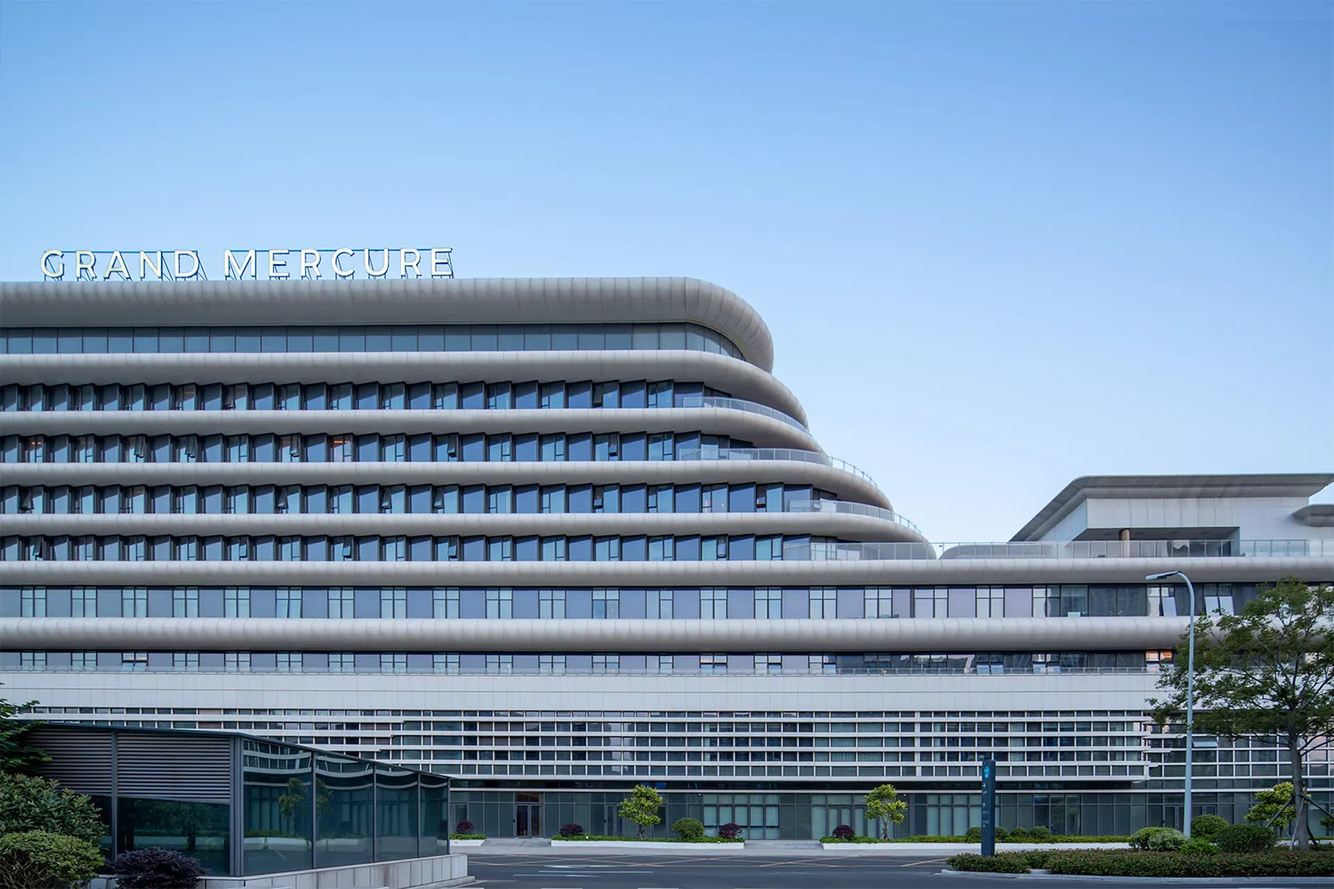 Exterior view of the Grand Mercure hotel featuring a modern curved, multi-layered white facade with extensive glass windows under a clear blue sky.