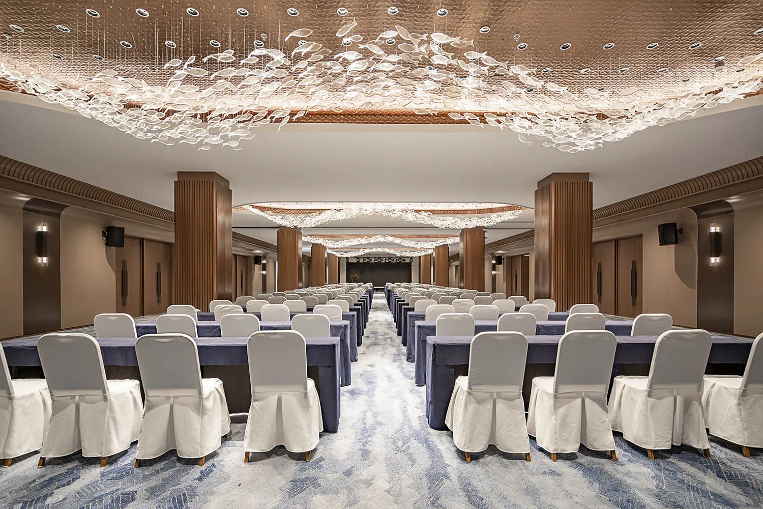 Luxurious grand ballroom prepared for an event with rows of tables covered in navy blue tablecloths and white chiavari chairs, dominated by a massive sculptural chandelier made of countless transparent acrylic fish hanging from a textured gold ceiling.