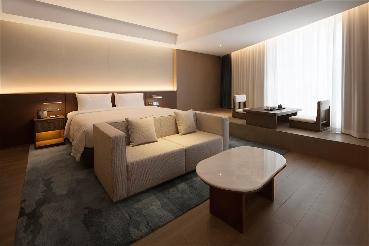 Modern luxury hotel bedroom featuring a large bed with backlit headboard, a beige sofa, marble coffee table, and a raised wooden platform with traditional tea seating beside a large window with sheer curtains.
