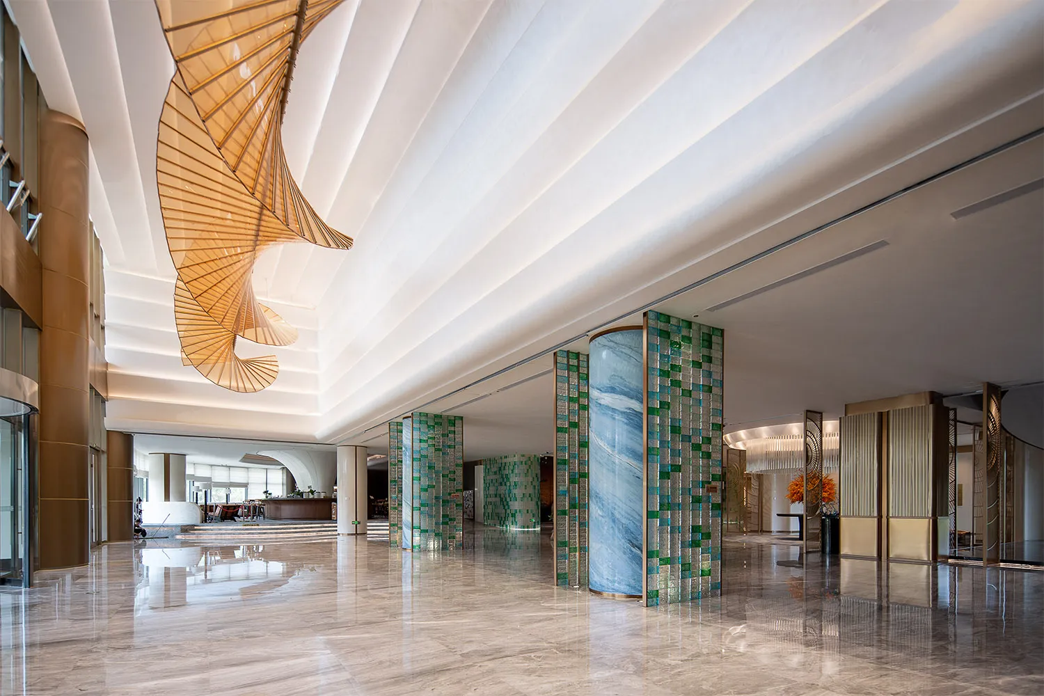 Spacious luxury hotel lobby with polished marble floors, blue and green mosaic pillars, and a large golden spiral chandelier hanging from a coffered ceiling.