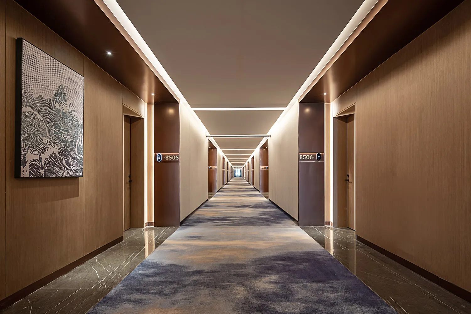 Long modern luxury hotel corridor with warm wood panel walls, dark marble flooring, patterned blue and beige carpet runner, recessed ceiling lighting, and large abstract mountain artwork on the left wall.