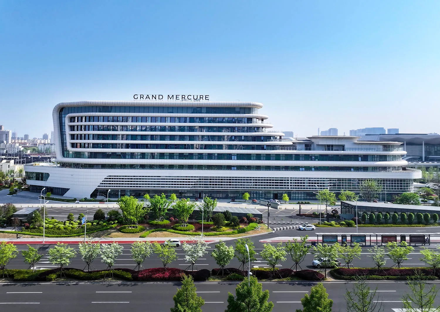 Front view of the modern Grand Mercure hotel building with distinctive curved white facade, extensive glass windows, and landscaped garden under a clear blue sky.