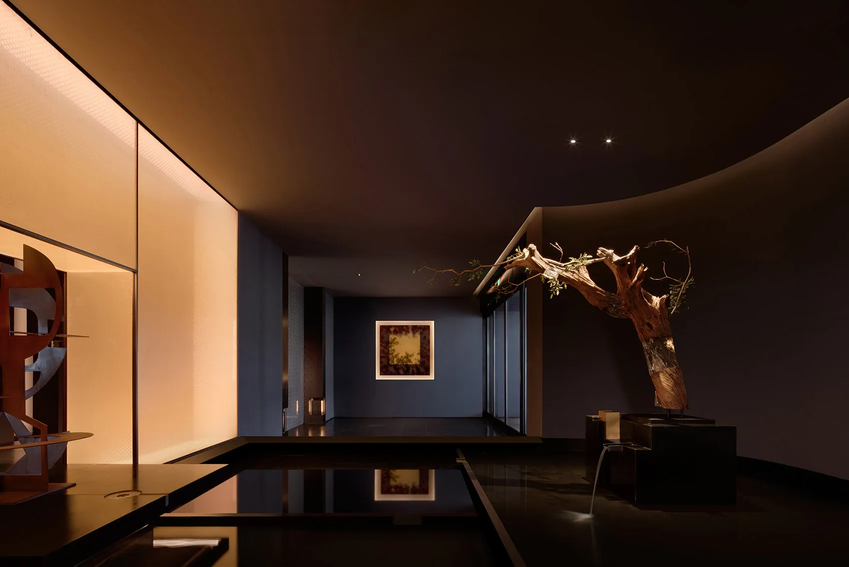 Modern dark hallway featuring a large sculptural dead tree installation, reflective black floor, curved ceiling, and warm backlit wall panels.