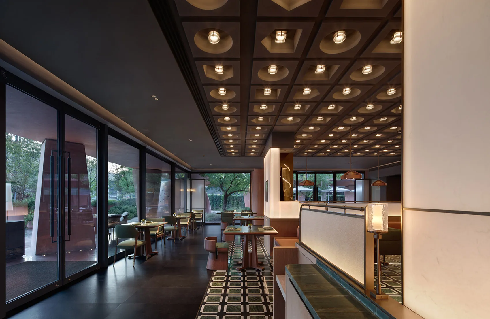 Modern elegant restaurant interior with dark coffered ceiling, recessed lighting, walnut dining tables, green upholstered chairs, and large glass windows overlooking greenery.