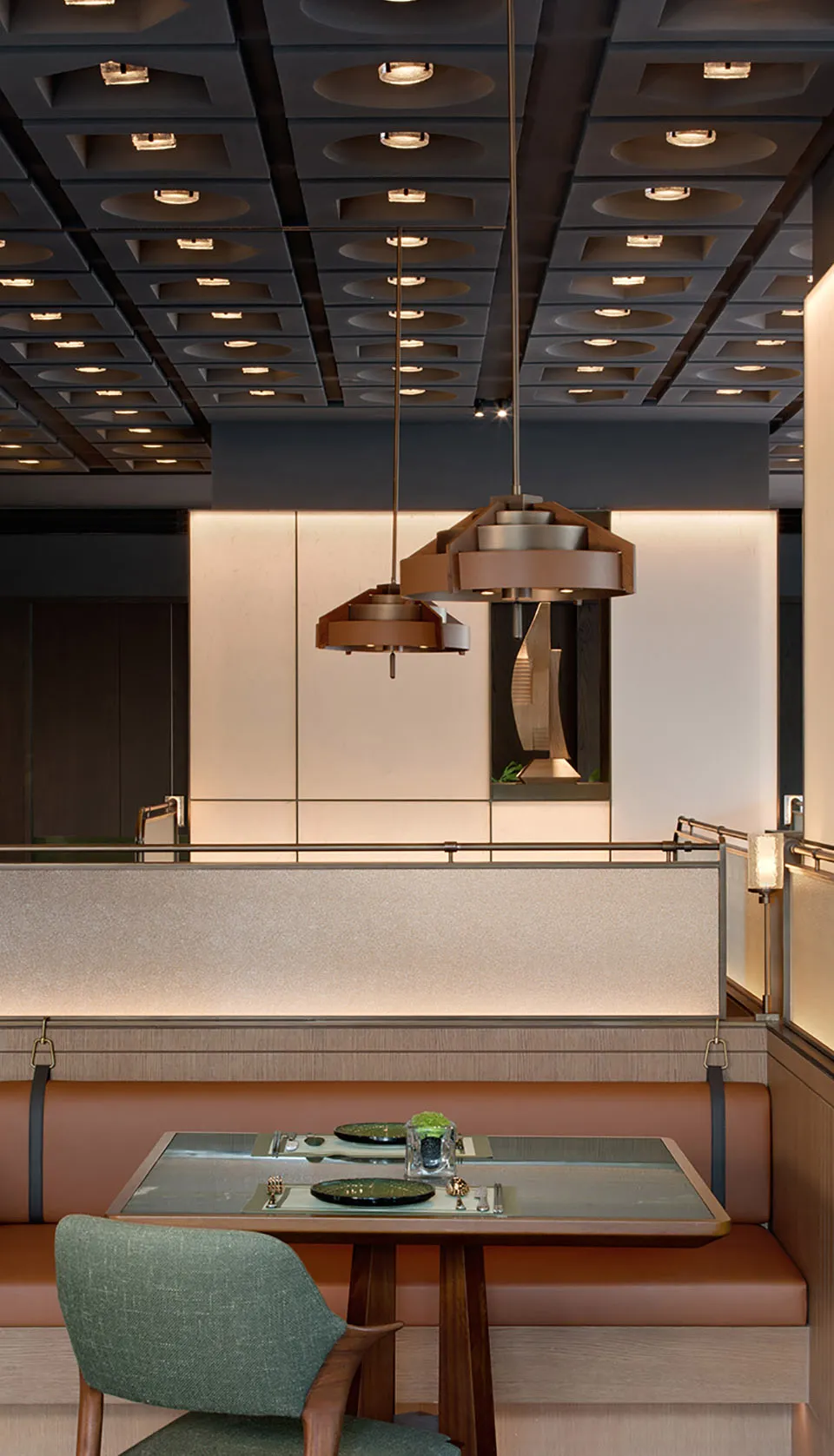 Modern restaurant dining booth with terracotta leather banquette, walnut dining table, green upholstered chair, and large geometric wooden pendant light hanging from a coffered ceiling.