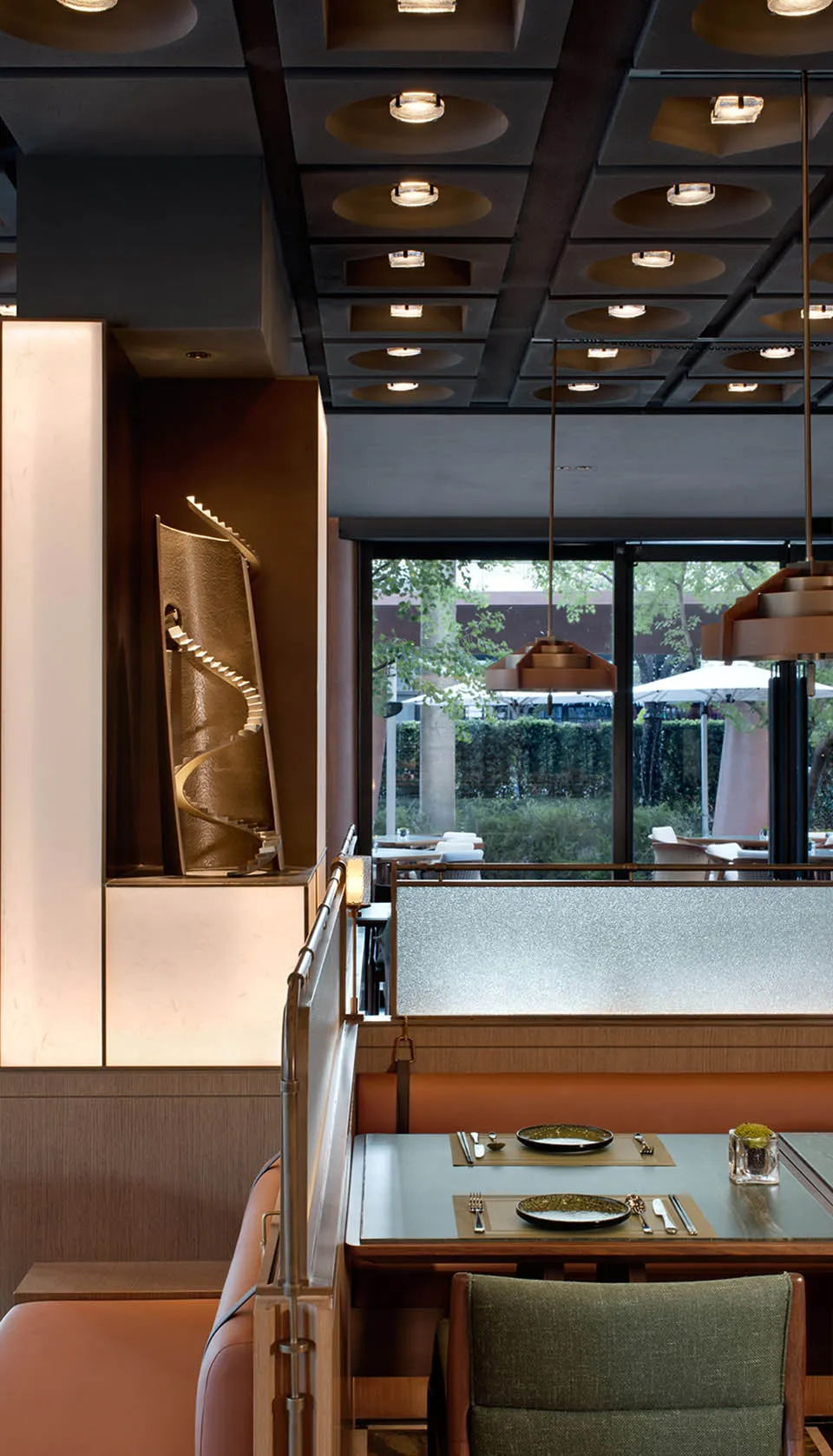 Elegant restaurant interior featuring a green upholstered dining chair, leather booth seating, a sculptural bronze spiral artwork, and dramatic ceiling lighting with large windows overlooking greenery.