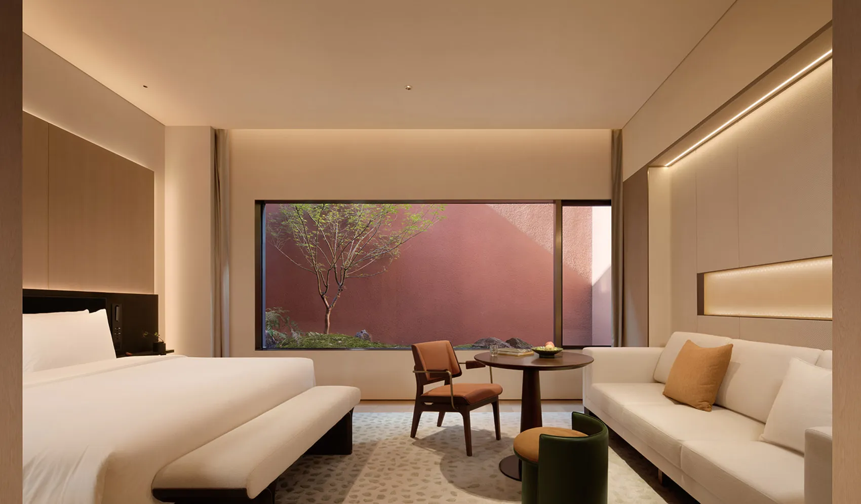 Modern luxury hotel room featuring a large bed with white linens, a white sofa, wooden armchair, round coffee table, and a large window overlooking a private Japanese-style garden with a red wall and small tree.