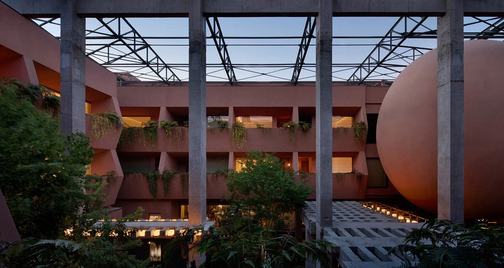 View of a brutalist architectural courtyard in dusty pink concrete with large geometric openings, hanging plants, and a massive spherical concrete sculpture at dusk.