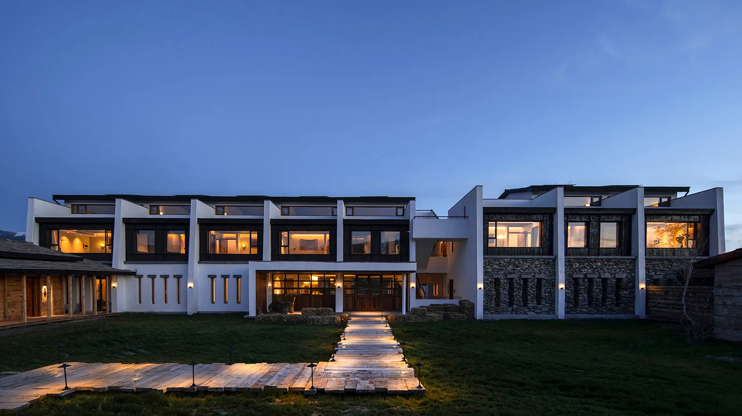 Modern farmhouse exterior at twilight featuring white walls, dark wood windows, stone cladding, and a glowing wooden boardwalk leading to the entrance.