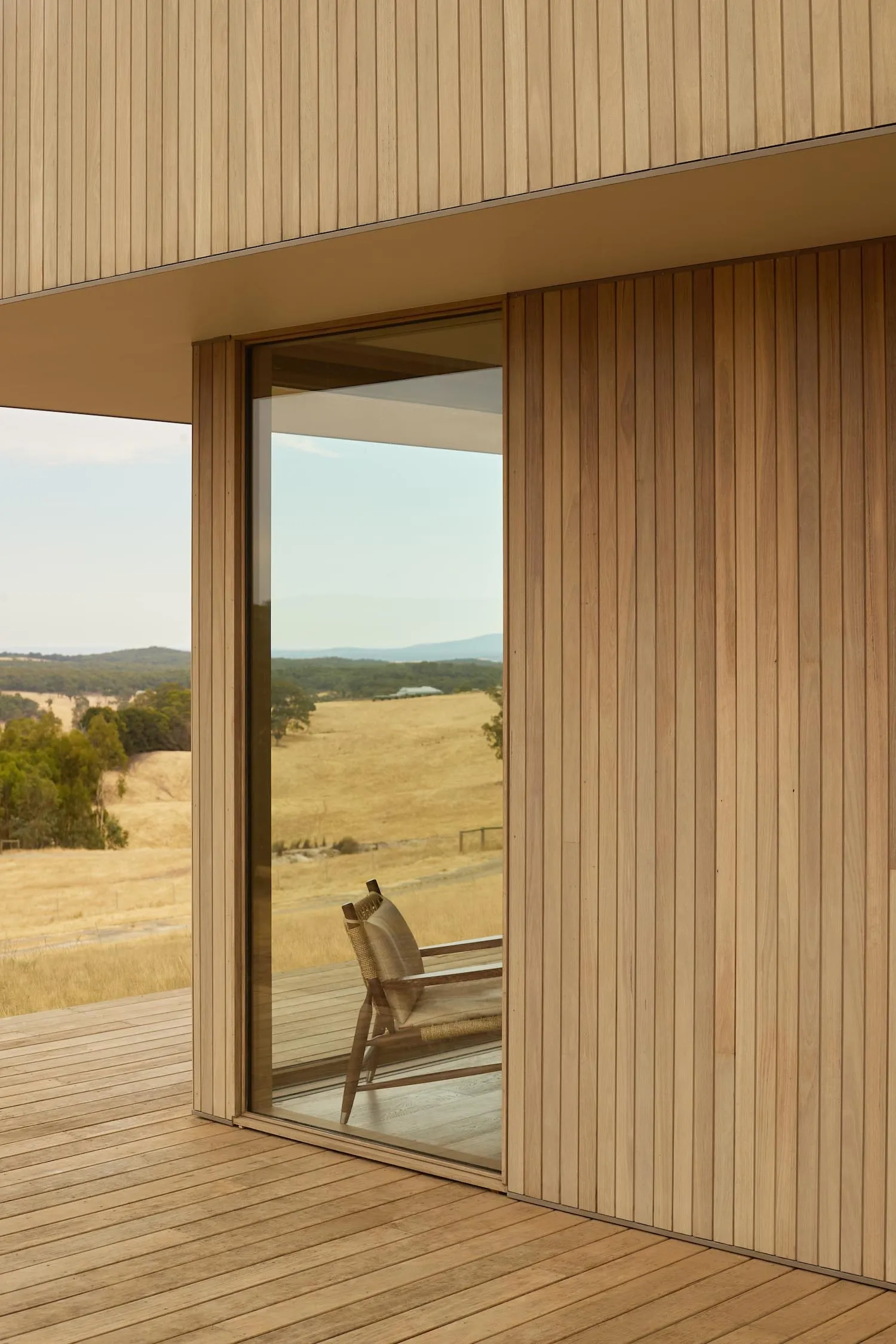 Modern house interior with vertical timber cladding opening onto a wooden deck overlooking golden grasslands and distant hills.