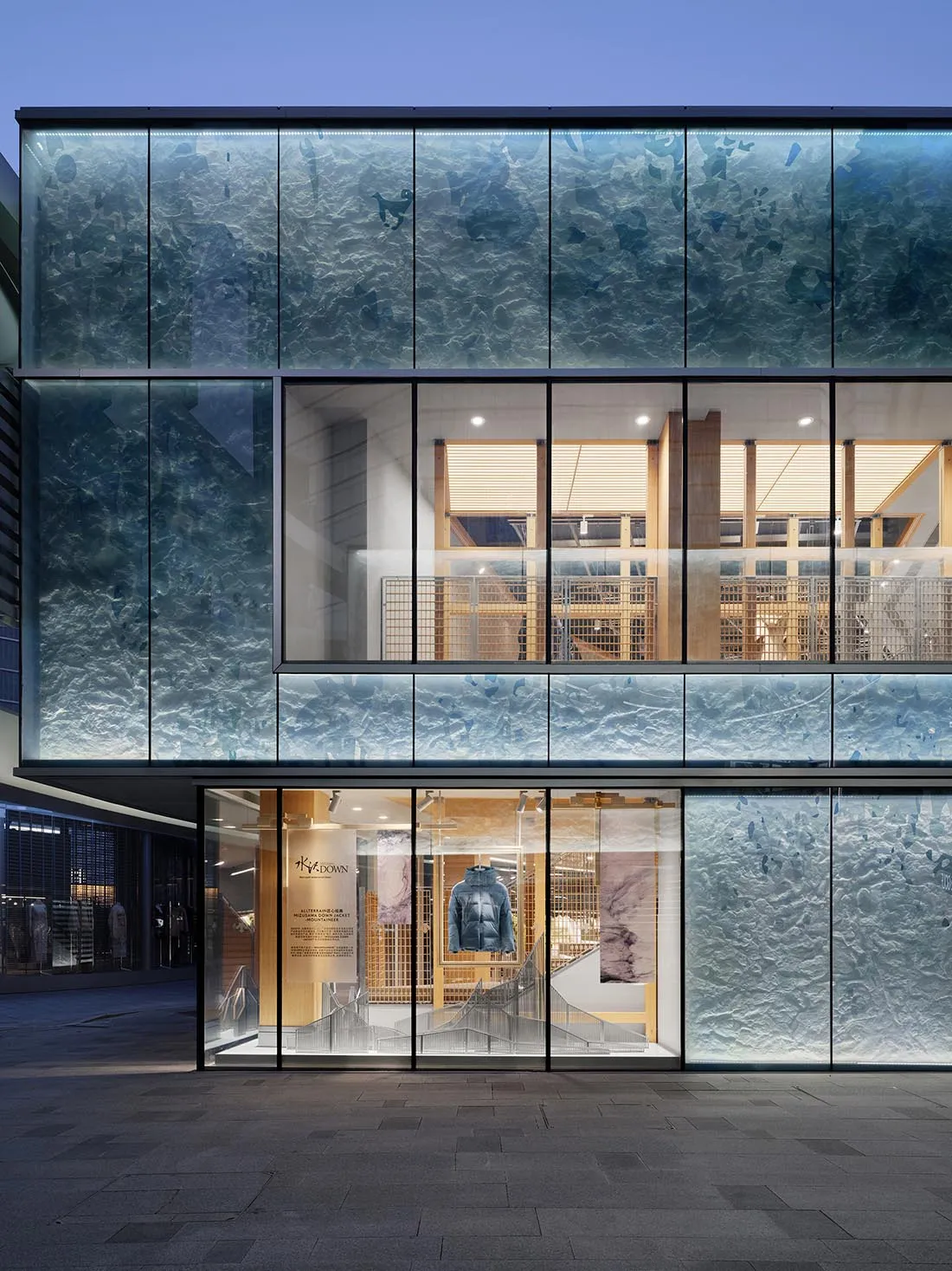 Modern retail storefront of K-DOWN featuring large textured blue glass panels resembling ocean waves, with warm wooden interior visible through the glass at night.