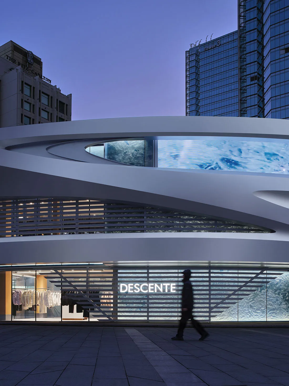 Night view of the modern curved facade of Descente store in Taipei with large digital ocean wave display and a pedestrian walking by.